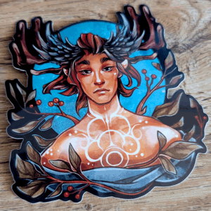 Photo of a sticker showing an androgynous figure with a white scrolling tattoo down their neck/chest. They have auburn hair and antlers that are framed by feathered crests